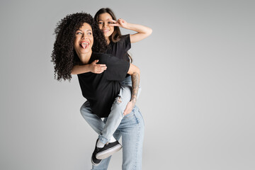 Positive and tattooed mother piggybacking preteen daughter showing peace sign isolated on grey.