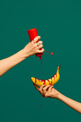 Female hand pouring ketchup at banana against green studio. Extraordinary taste. Food pop art...