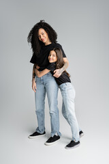 Full length of happy mother and daughter in t-shirts and jeans hugging on grey background.