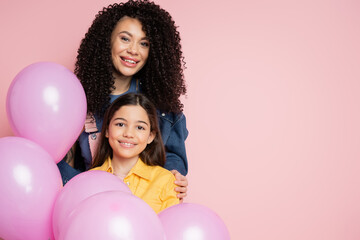 Positive woman looking at camera near daughter and party balloons isolated on pink.