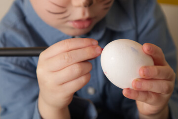 The hands of a little boy hold a chicken egg and a felt-tip pen and paint an easter egg close up