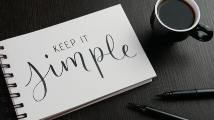 KEEP IT SIMPLE lettering in notebook with cup of coffee and pens on black wooden desk