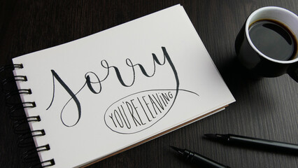 SORRY YOU'RE LEAVING lettering in notebook with cup of espresso and pens on black wooden desk