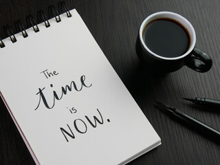 THE TIME IS NOW lettering in notebook with cup of coffee and pens on black wooden desk