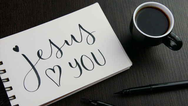 JESUS LOVES YOU with heart symbol lettering in notebook with cup of espress and pens on black wooden desk