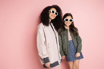 Positive mom and stylish daughter in sunglasses holding hands on pink background.