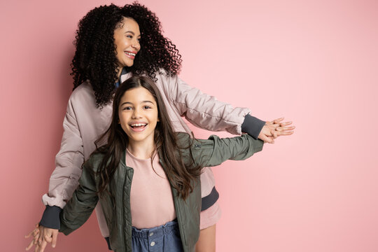 Positive curly woman holding hand of preteen daughter on pink background.