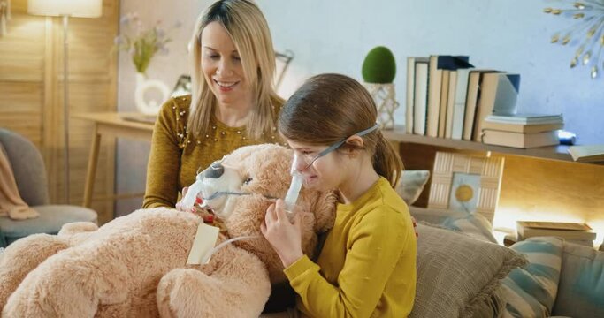 Mother and daughter at home playing with a teddy bear while the girl inhales using an inhaler