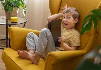 A little boy sits in a chair with the book and communicates with a smart speaker