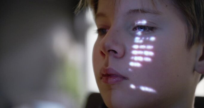 Closeup shot of a young boy sitting in front of a computer and scrolling thru text. On his face is a projection from the pc.