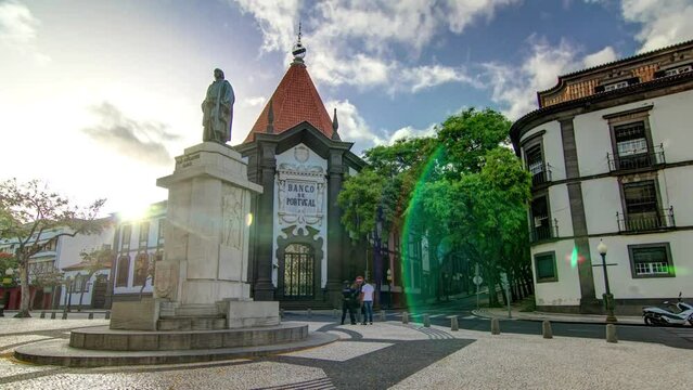 A statue of Zarco stands on the Avenida Arriaga timelapse hyperlapse and Banco de Portugal in the background in Funchal, Madeira, Portugal. Green trees and clouds on the sky