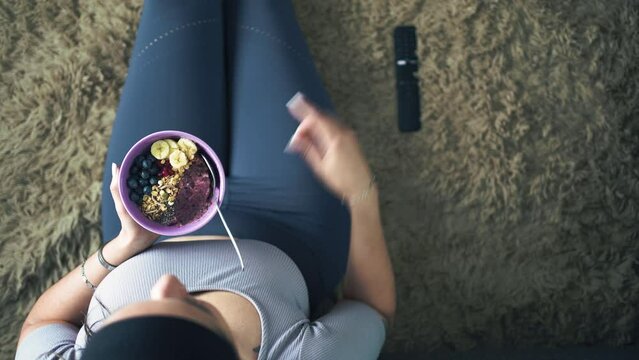 Unrecognizable oman wearing sport outfit eating healthy granola and smoothie bowl while watching television sitting on the floor. Top view video shot