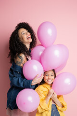 Excited woman standing near daughter and balloons on pink background.