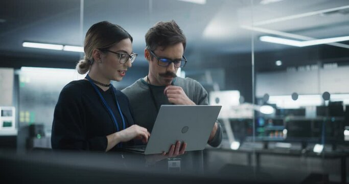 Portrait of Two Creative Young Female and Male Engineers Using Laptop Computer to Analyze and Discuss How to Proceed with the Artificial Intelligence Software. Standing in High Tech Research Office