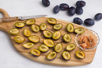 Wooden board with chopped and peeled plums, halves of plums, a bowl with pits, whole plums and a kitchen knife on a light gray background, top view. Making homemade jam or plum cake