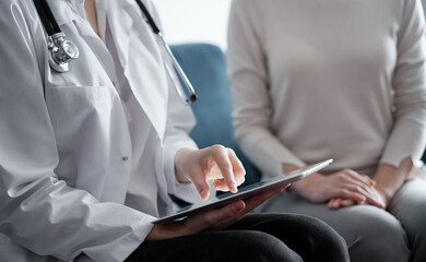 Doctor and patient sitting at sofa in clinic office. The focus is on female physician's hands using tablet computer, close up. Perfect medical service and medicine concept.
