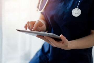 Obraz premium Doctor woman using tablet computer while standing near panorama window in clinic, close up. Physician or surgeon at work. Medicine concept.