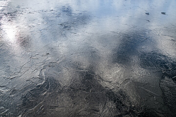 The texture of thin melting ice on a spring lake with the sky reflected on the surface. Light glare.