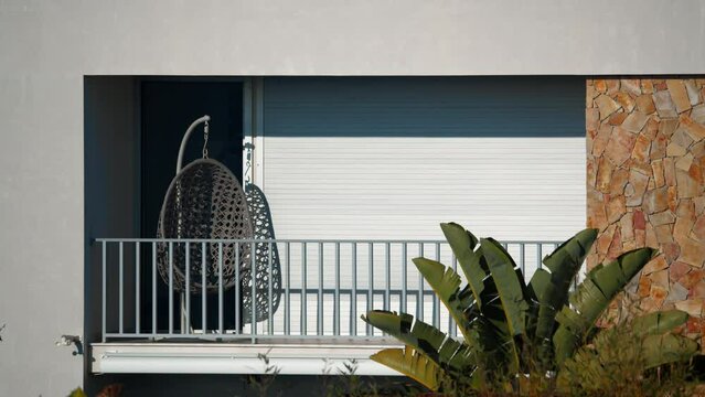 A Balcony With A Swing Chair Covered By Closed Blinds, Surrounded By Lush Vegetation And Bright Sunlight, With No People In The Frame. Filmed In Portugal