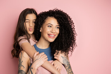 Preteen kid hugging tattooed mom and pouting lips on pink background.