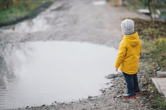 A child in a yellow raincoat and rubber boots runs through the puddles. Little boy plays in the water 2-3 years old