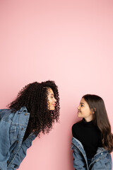 Side view of trendy mother and child in denim jackets standing on pink background.