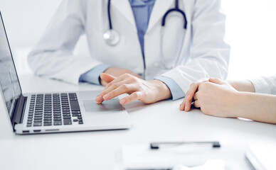 Fototapeta premium Doctor and patient sitting near each other at the desk in clinic. The focus is on female physician's hands using laptop computer, close up. Medicine concept.