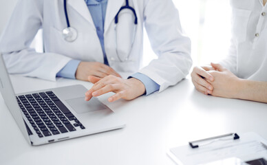 Doctor and patient sitting near each other at the desk in clinic. The focus is on female physician's hands using laptop computer, close up. Medicine concept.