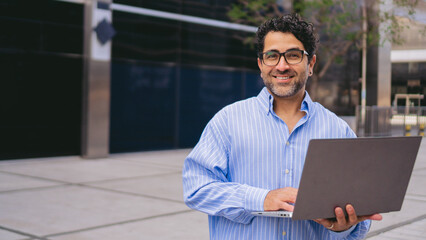 Panoramic photo of a middle-aged latin man standing looking at camera with a laptop in his hands. Copy space.