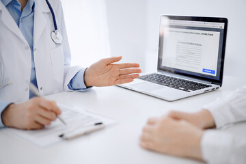 Fototapeta premium Doctor and patient sitting opposite each other at the desk in clinic. The focus is on female physician's hands pointing into laptop computer monitor, close up. Medicine concept.