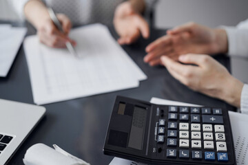 Woman accountant using a pen and laptop computer while counting and discussing taxes with a client, focus on the calculator. Business audit and finance concepts.