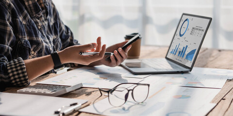 Young accountant hand holding smartphone to call marketing consultants and using laptop computer to...