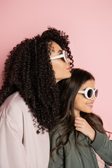 Curly woman in sunglasses standing near daughter in bomber jacket on pink background.