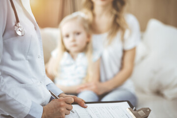 Doctor and patient. Pediatrician using clipboard while examining little girl with her mother at home. Sick and unhappy child at medical exam