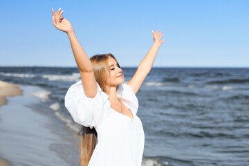 Fototapeta na wymiar Happy, beautiful woman on the ocean beach standing in a white summer dress, raising hands.