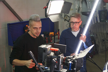 two caucasian male videographer and light gaffer install studio lighting on the set and watch the result on the monitor screen of a professional movie camera