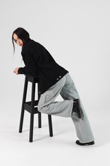 Portrait of a beautiful brunette girl leaned on a bar chair, dressed in a black jacket, wide jeans. Isolated on white.