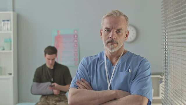 Medium portrait of mid aged Caucasian military doctor in scrubs posing for camera with hands folded in bright modern office while young soldier with broken arm sitting on medical couch in background