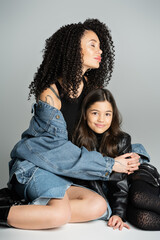 tattooed and curly woman hugging happy child while sitting on grey background.