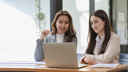 Two beautiful Asian businesswoman in conversation Exchange ideas at work Company employees working together by talking and guiding each other with papers, graphs and desk laptops.
