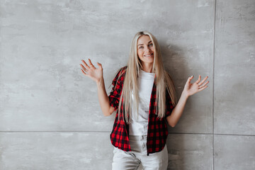 Adorable blonde Scandinavian young woman with long hair in plaid shirt and white pants spreads hands stands at marble wall with copy space. Repair, cozy home. Satisfied Italian girl home