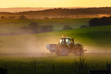 Fototapeta premium Tractor in farmland working on soil field with sunset, big equipment, countryside scene
