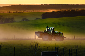 Fototapeta premium Tractor in farmland working on soil field with sunset, big equipment, countryside scene