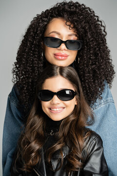 Portrait of curly woman in sunglasses posing near smiling daughter isolated on grey.