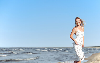 Fototapeta na wymiar Happy blonde woman in free happiness bliss on ocean beach standing straight. Portrait of a female model in white summer dress enjoying nature during travel holidays vacation outdoors