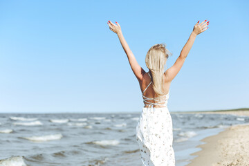 Happy blonde beautiful woman on the ocean beach standing in a white summer dress and sun glasses, raising hands.