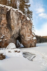 Fototapeta na wymiar Winter landscape with a rock in the form of a horse 's head drinking water and trees on the bank of a snow - covered river against a cloudy blue sky