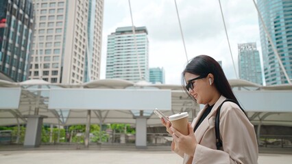 Asian business women going to work in the morning with skyscrapers, She was smiling in her hand holding smartphone and paper cup of coffee