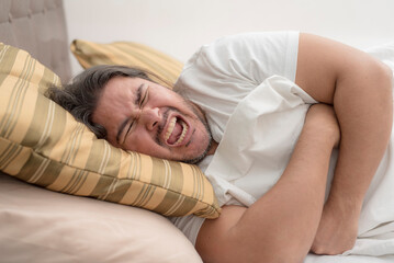 Obraz na płótnie Canvas A man screams while clutching his ribcage while resting on his side on the bed. Possible pulled muscle or rib fracture.