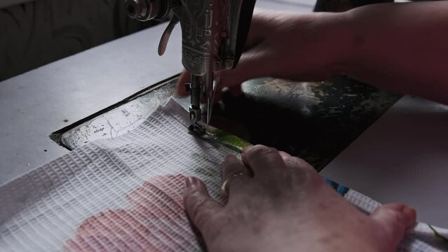 An aged seamstress sews at a traditional sewing machine at home in slow motion. Close-up of aged female hands working at a retro sewing machine. The needle quickly moves up and down. Lifestyle, hobby